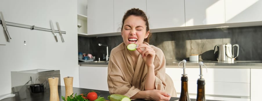 Portrait of happy girl eating vegetables while making meal, cooking healthy vegetarian food in kitchen, holding zucchini, posing in bathrobe.