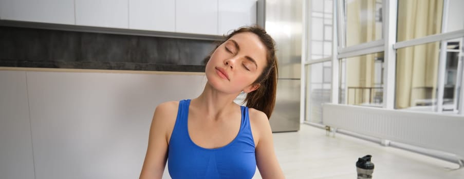 Portrait of young sportswoman, stretching her neck, warm-up before yoga exercises, doing fitness workout on rubber mat, wearing blue sportsbra and leggings.