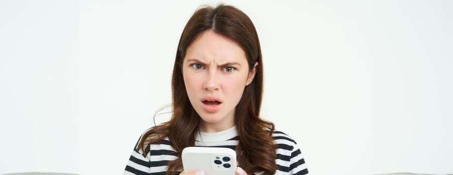 Portrait of woman holding smartphone, looking offended and confused at camera after reading message on mobile phone, white background.