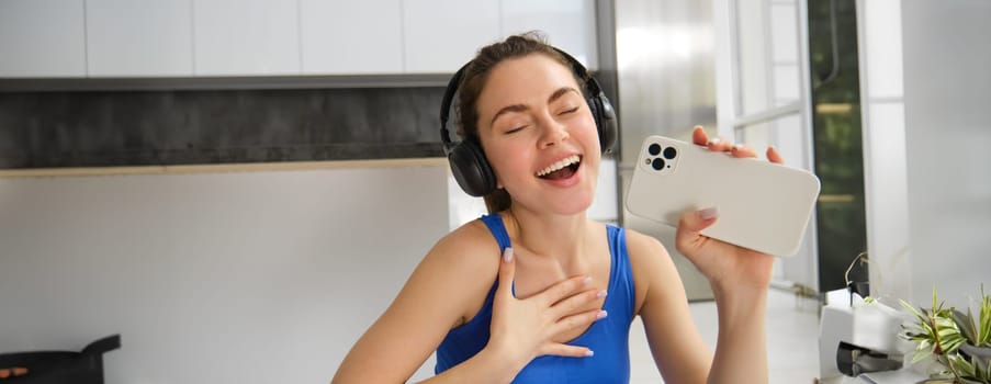 Close up portrait of young happy woman, workout at home, listens music in wireless headphones, singing at smartphone, doing training exercises.