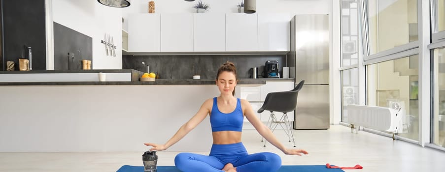 Portrait of calm and relaxed young woman, sitting on yoga rabber mat, concentrating on breathing exercises, doing mindful training at home. Copy space