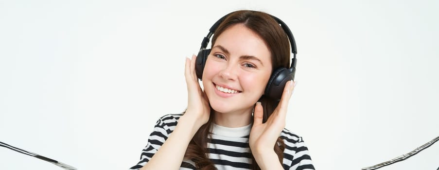 Portrait of beautiful woman in wireless headphones, listening music, using earphones, smiling at camera, standing over white background.