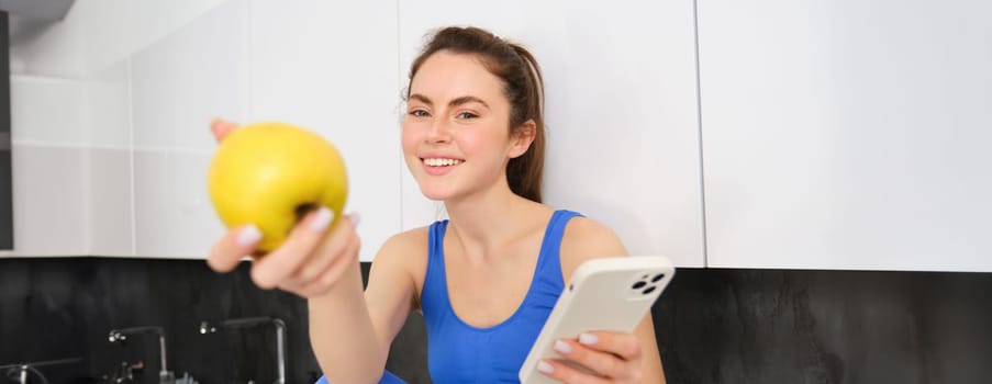Image of beautiful, sportswoman sitting in kitchen with smartphone, offering you an apple, eating fruits, wearing sportsbra and leggings.
