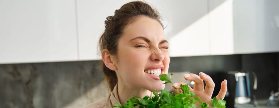 Funny and cute brunette woman bites delicious, fresh green parsley, eating herbs, cooking salad in kitchen.