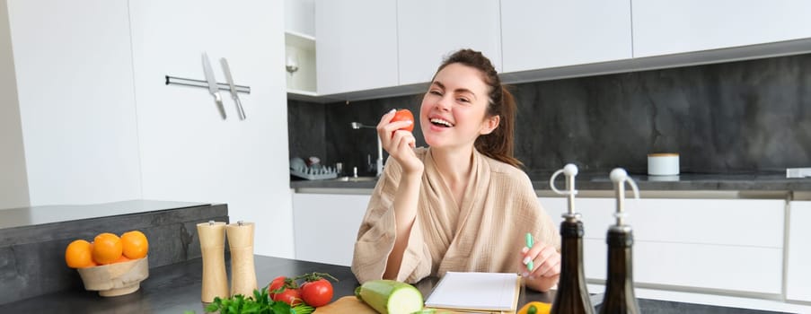 Portrait of happy brunette woman, bites tomato, sits in the kitchen with vegetables, cooking vegetarian meal, preparing list of groceries, dinner menu, wearing bathrobe.