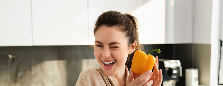 Close up portrait of cute winking girl, showing yellow pepper, standing in kitchen, making vegetarian meal, making salad, eating healthy vegetables.
