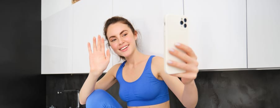 Portrait of sportswoman, fitness blogger, recording video, waving hand at smartphone screen and smiling, online chatting, sitting in kitchen, wearing activewear.