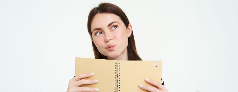 Portrait of young woman makes notes in her planner, writes in diary, checks her schedule in notebook, holding organizer in hands, stands over white background.