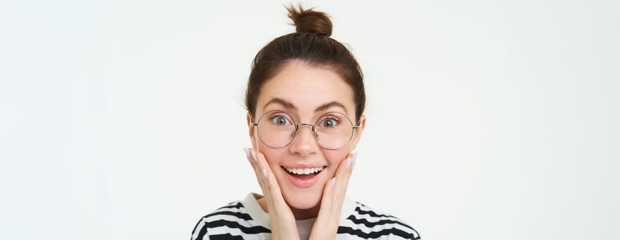 Image of amazed, smiling young woman in eyewear, wearing glasses, looking impressed and excited, standing over white background.