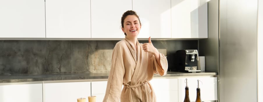 Portrait of happy young woman preparing vegetarian dinner, showing thumbs up, wife cooking in kitchen and smiling, recommending product, chopping vegetables for healthy salad.