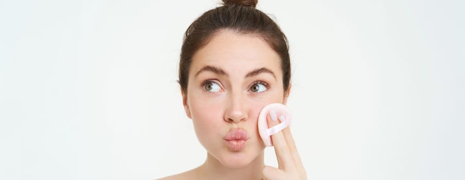 Portrait of woman washing off her face, takes off makeup with cosmetic pad, puts on skincare cleanser, stands over white background.
