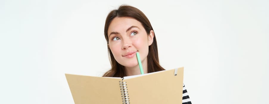 Portrait of creative young woman, looking thoughtful, holding pen and notebook, writing in her planner, thinking while making notes, standing over white background.