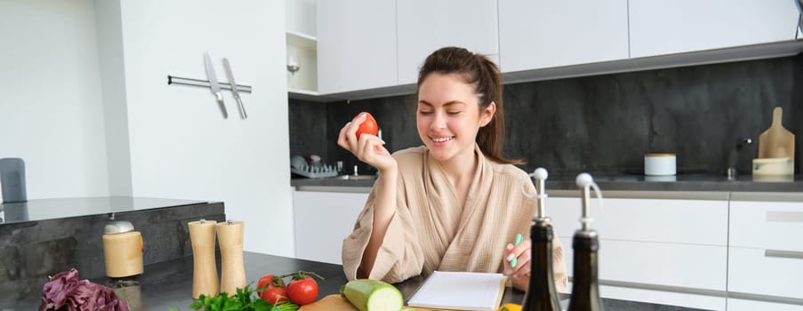 Portrait of beautiful young woman in the kitchen, writing down cooking recipe, sitting near chopping board with vegetables and making grocery list, creating healthy veggie menu for her family.