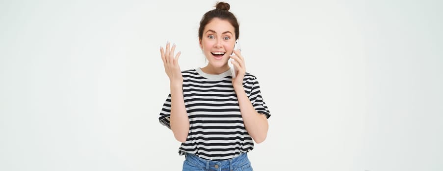 Portrait of excited woman answers phone call, reacts amazed to wonderful news received over the telephone, standing over white background.