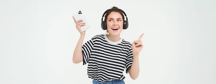 Portrait of happy brunette woman, dancing in headphones, listening music on her mobile app, enjoys her favorite song, isolated against white background.