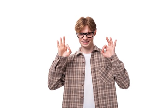 young stylish european student man with golden hair in glasses and a plaid shirt shows ok.