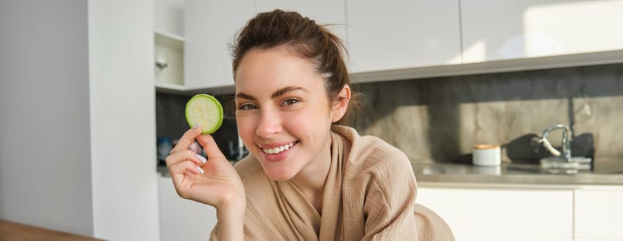 Portrait of happy, smiling young woman in the kitchen, cooking, chopping zucchini, holding vegetables and looking happy, preparing vegan food meal at home.