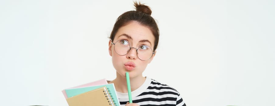 Close up portrait of girl in glasses, thinking, holding notebook and pen, looking up and pondering, making decision, standing over white background.