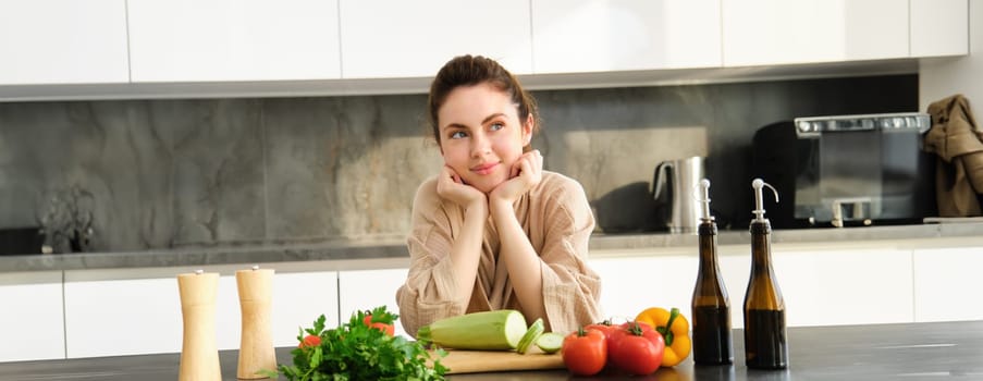 Portrait of smiling young woman leaning on kitchen counter, preparing dinner, standing near chopping board, cutting zucchini, looking away and thinking, thinks what to cook.