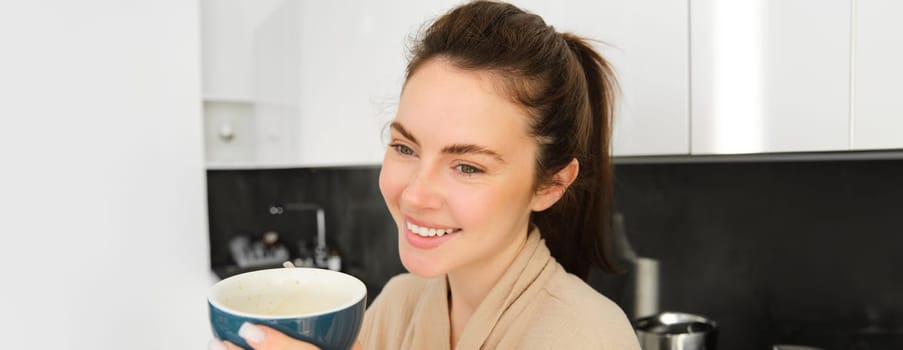 Portrait of good-looking young woman starting her day with cup of coffee, standing in the kitchen and drinking cappuccino from big mug, enjoying favourite drink in the morning.