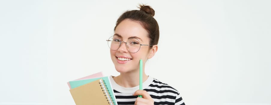 Portrait of smart girl in glasses, tutor holding pen and notebooks, student carry her homework notes, standing over white background.