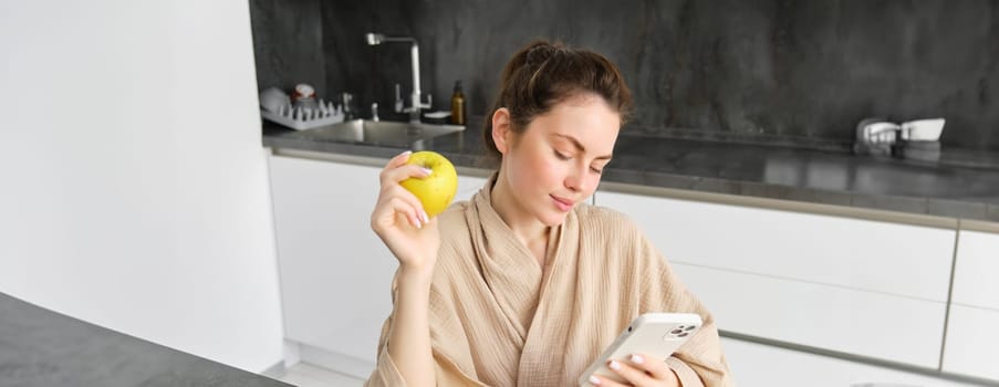Image of attractive young woman in bathrobe, sits in kitchen, eats green apple and looks at mobile phone, uses smartphone app, orders food on application.