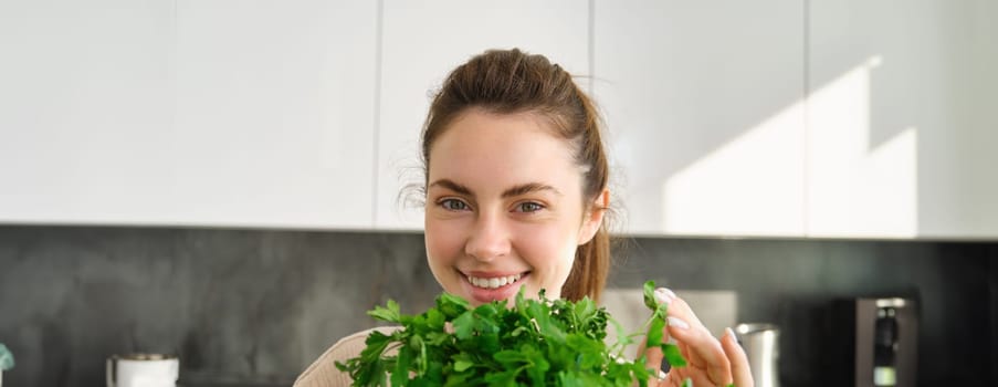 Portrait of beautiful smiling girl with bouquet of parsley, standing in the kitchen and cooking, adding herbs to healthy fresh salad or meal, preparing food.