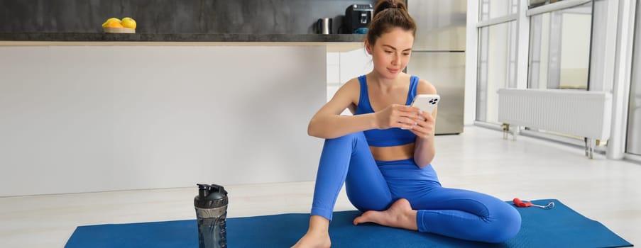 Portrait of young fitness girl sits on yoga mat with water bottle, looks at smartphone app, follows yoga training instructions.