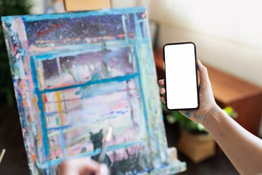 Designer or Artist holding blank screen smartphone over canvas of an artist with art.