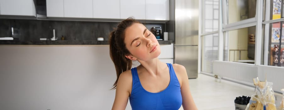Close up portrait of young woman stretching her neck, doing yoga exercises, fitness training at home, meditating in living room.