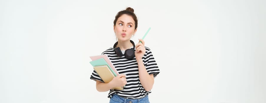 Portrait of young woman standing with books, student looking thoughtful at copy space, thinking about something, holding notebooks and homework in hands, white background.
