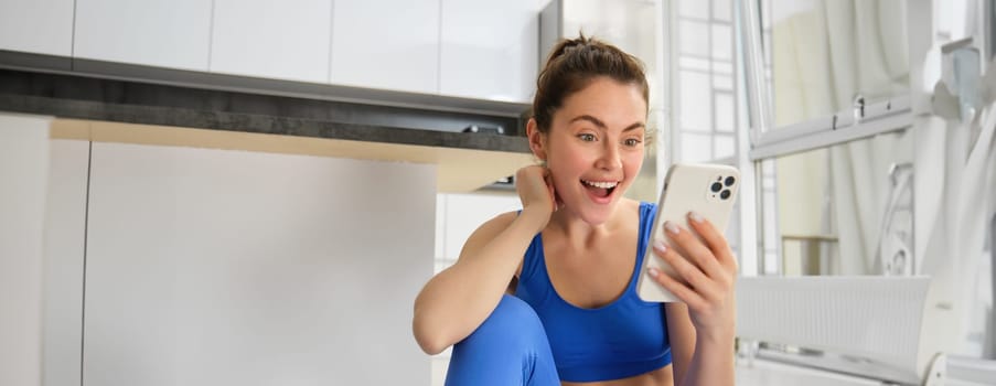 Close up shot of young happy woman, doing sports, workout at home, checking her phone, looking amazed at smartphone screen.