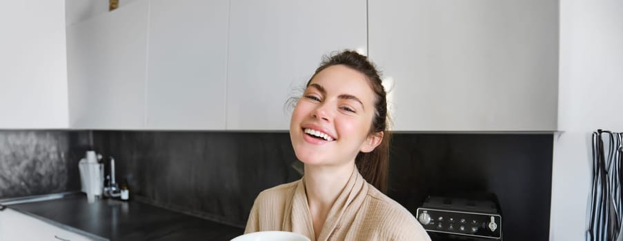 Lifestyle concept. Portrait of happy brunette woman in bathrobe, drinking coffee in the kitchen, having morning cuppa and smiling.
