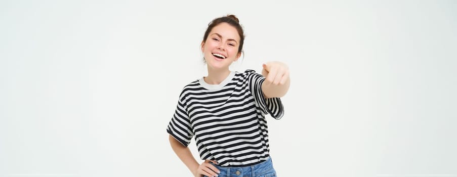 Image of stylish young woman, pointing finger at camera, inviting you, standing over white background.