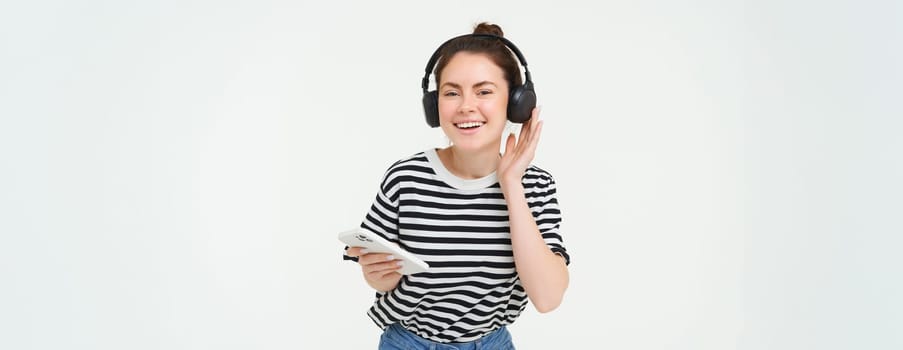 Portrait of happy woman with smartphone changes song on mobile phone streaming app, listens music in headphones, dancing against white background.