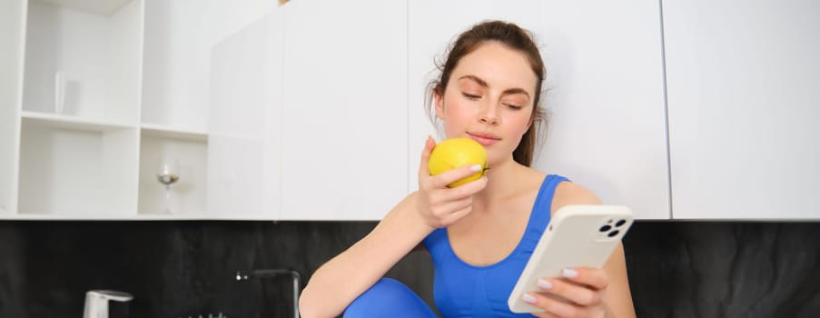 Image of stylish sportswoman, young fitness instructor sitting in kitchen and eating an apple, holding smartphone, using social media app on mobile phone.