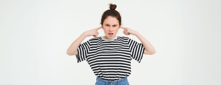 Image of annoyed, bothered woman plugs, shuts her ears with fingers, disturbed by loud noise, stands over white background.