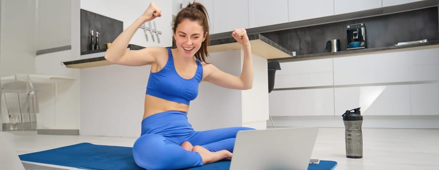 Image of excited young woman showing muscles, flexing biceps and looking at laptop screen, workout from home, fitness instructor teaching remotely, giving online yoga lesson.