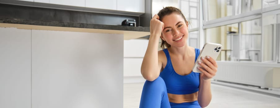 Young woman with smartphone does workout at home, using mobile phone app for sports training indoors, wears blue sportsbra and leggings.