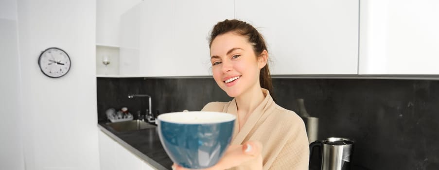 Portrait of good-looking young woman gives you cup of coffee, offering mug to you, standing in the kitchen, wearing bathrobe.