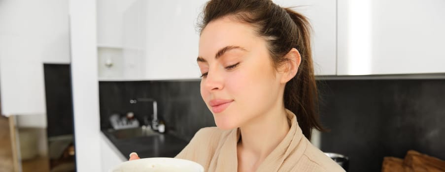 Close up portrait of attractive girl drinking coffee, holding cup with morning cappuccino and smiling, having a mug of delicious drink in the kitchen, wearing comfortable bathrobe.