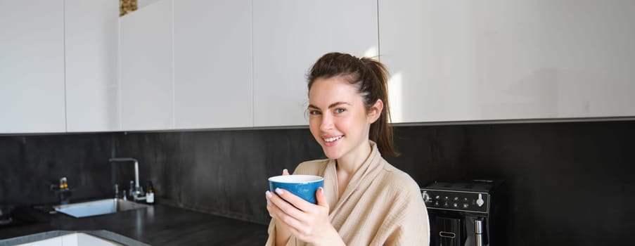 Portrait of carefree smiling woman drinking coffee, standing in kitchen with delighted, pleased face expression, waking up in the morning and having cappuccino.