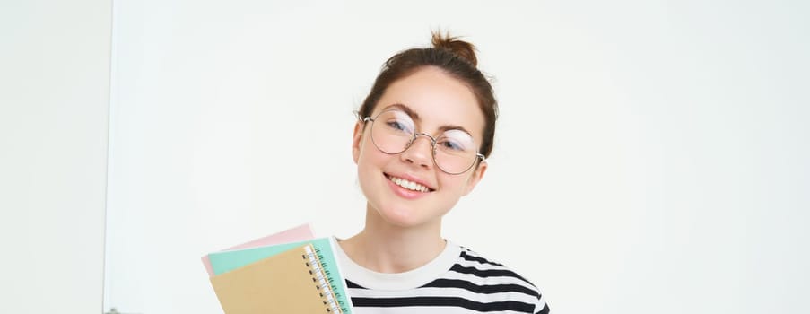 Portrait of young woman, tutor in glasses, holding her notebooks and documents, smiling at camera, standing over white background.