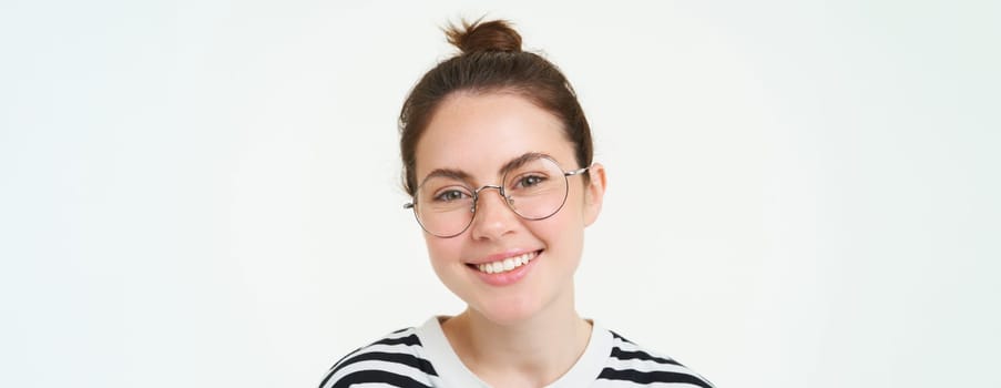 Close up portrait of cute young woman in glasses, looking at camera and smiling, trying on new spectacles, standing over white background.