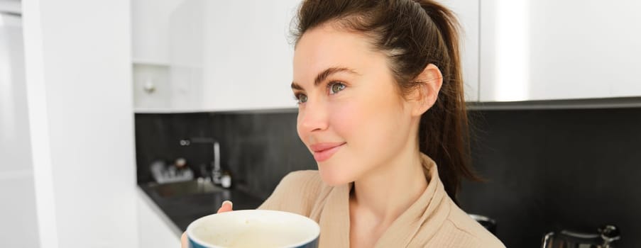 Portrait of beautiful young woman in bathrobe, drinking morning coffee and enjoying the taste, smiling pleased, standing in the kitchen.