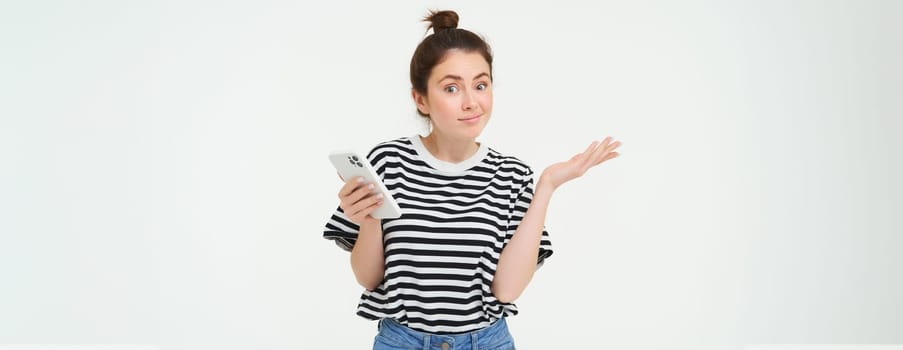 Image of clueless girl shrugs shoulders with smartphone in hand, isolated over white background.