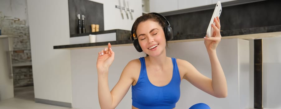 Cheerful brunette girl does fitness at home, wears wireless headphones and dances, holds smartphone, does workout indoors on floor, wears blue activewear.