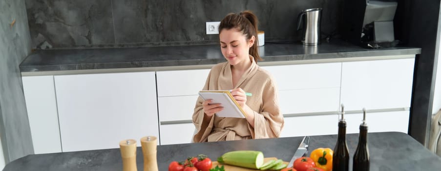 Portrait of happy young woman writes down menu for dinner, sits in the kitchen near vegetables, makes grocery list for shopping, poses in the bathrobe.