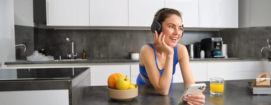 Image of happy, stylish young sports woman, standing in kitchen and drinking orange juice, listening music in headphones, using smartphone app.