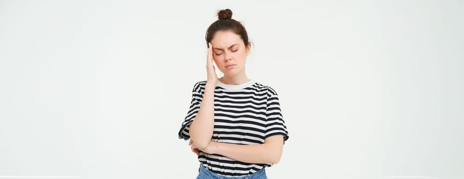 Portrait of tired woman rubs her forehead, touches head, has a headache, stands distressed against white background.